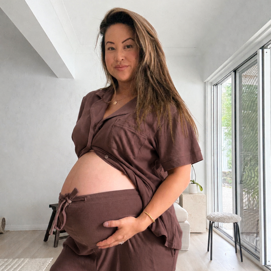 Pregnant woman standing in a bright room wearing chocolate maternity pyjamas open over her bare bump