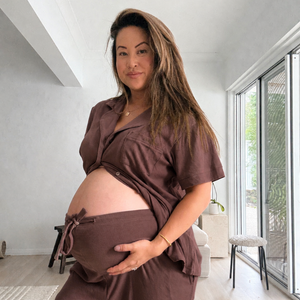Pregnant woman standing in a bright room wearing chocolate maternity pyjamas open over her bare bump